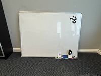 Photo of large whiteboard with magnets, markers, eraser, and cleaning supplies on carpeted floor against wall.