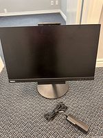 Front view of Lenovo ThinkCentre Tiny-in-One monitor with round stand and cables laid in front on carpeted floor.