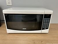 Closed view of white RCA microwave on wooden surface showing front including black glass door and control panel.