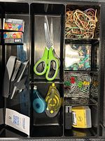 Photo of black compartmentalized tray filled with various office supplies including scissors, staplers, correction tape, staples, rubber bands, push pins, and paper clips.