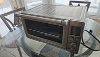 Full view of the stainless steel Breville toaster oven on a table, showing the door, handle, and power cord.
