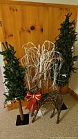 Two pre-lit artificial Christmas trees, white decorative branch arrangement with large red and gold bow, and a wicker reindeer decoration, all arranged grouped against a wood paneled wall.