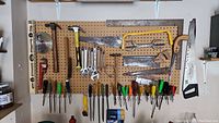 Photo of assorted hand tools hanging on a wooden pegboard showing saws, hammers, wrenches, screwdrivers, and a pipe wrench.