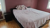 King size bed frame with extra deep pillow top mattress covered in light bedding in room with wood floor and pink walls