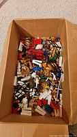Top-down view of a cardboard box filled with assorted toy figures and Lego pieces including horses and small fencing parts.