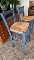 Two blue painted wooden bar stools with woven straw seats shown side by side on tile floor beside cabinetry.