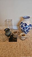 Photo showing a large ceramic vase with blue floral pattern, two small striped pottery containers, two clear glass vases, and a small dark statue on a base, arranged on carpet.