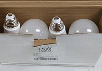 View of four Noger 15W LED light bulbs inside partially open white cardboard box showing bulb shape and screw base.