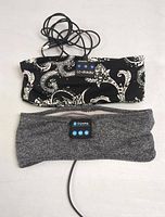 Two Bluetooth headbands side by side, one black paisley patterned and one gray, both showing control panels and attached USB charging cables.