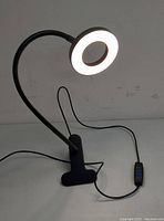Clip-on LED ring light switched on, showing bright white circular light with flexible arm and attached clip, power cord visible with inline remote control.