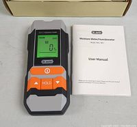 Digital moisture meter with backlit screen showing 0% moisture reading, user manual beside it, white background