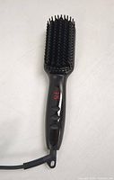 Front view of electric hair brush showing paddle bristles, ergonomic black handle with three buttons and cord attachment.