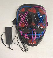 Front view of a black Halloween mask with pink and blue LED lights forming X shapes over the eyes and stitched mouth design, plugged into a wired black battery pack with power button.