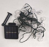 Photo showing LED solar string lights with multiple clear textured ball globes on green wiring, alongside attached solar panel with stake for outdoor placement.