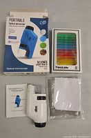 Photo showing the microscope, instruction manual, blue carrying case and the box of 12 prepared slides on a light surface.