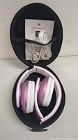 Purple Bluetooth over-ear headphones folded inside a black zippered carrying case with white USB cord, auxiliary cable, and user manual in case mesh pocket.