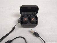 Sennheiser earbuds placed inside an open charging case with visible Sennheiser logo on earbuds, charging case lid open, USB charging cable partially visible.