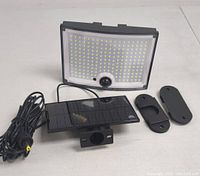 Outdoor solar LED flood light with motion sensor, detachable solar panel, and two mounting brackets arranged on a plain surface.
