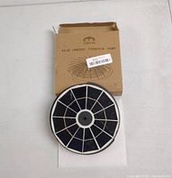 Solar powered fountain pump circular panel with black cells and white border, next to slightly worn brown cardboard box with product name and water spray graphic.