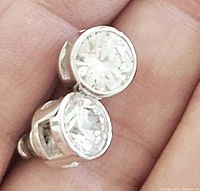 Close-up view of two round clear cubic zirconia stones set in silver bezel stud earrings held in hand.