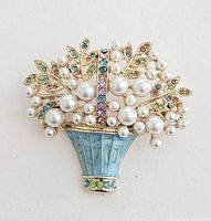 Front view of brooch showing faux pearls, multi-colored rhinestones, gold-tone metal basket with blue enamel