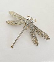 Gold tone dragonfly brooch with clear rhinestones arranged on wings and body shown on white background.