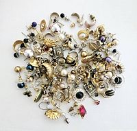 Top-down view of a large assortment of pierced earrings in various styles, colors, and materials including metallics, faux pearls, rhinestones, and beads spread on a white background.