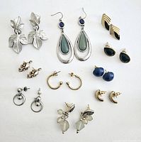 Photo showing all 10 pairs of pierced earrings arranged neatly on a white surface including silver leaf shapes, teardrop with blue and green stones, black and gold enamel, rhinestone clusters, gold hoops, blue beads, and crescent shapes.