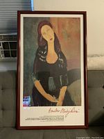 Framed poster of Amedeo Modigliani artwork depicting a modernist portrait of a woman with elongated features, dark hair, and seated pose. Poster includes exhibition details at bottom and artist signature in red ink style font.