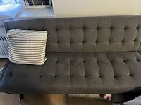 Gray tufted futon sofa against a wall with a single white and black striped cushion on left side. The sofa is in good condition and measures about 6 feet long.