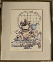 Framed watercolor print of teddy bear sitting on wicker chair with blue pillows.