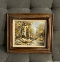 Front view of signed landscape painting in large brown wooden frame, showing trees and a stream in fall colors.