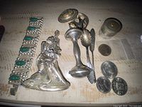 Full view of the lot: silver figurine, candlestick holder, medallion holder with lid removed showing wristwatch, silver coins, and bracelet.