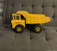 Photo showing large yellow metal Tonka dump truck on grey tufted surface, viewed from side showing details of dump bed, cab, and wheels.