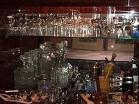 Wide shot showing two glass shelves with various clear glass drinking glasses, shot glasses, several decanters, metal bar tools including bottle openers and corkscrews, and wooden pestles.