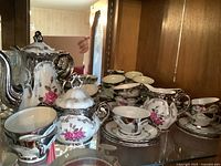 Multiple views of Bavaria porcelain demitasse coffee set showing teapot, sugar bowl, creamer, cups and saucers with pink rose and silver decoration.