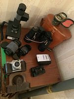 Full view of the lot on a wooden table showing Sunpak flash, multiple lenses, Zenith binoculars, goggles, and cases.