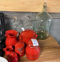 Photo showing red ceramic canisters, red ceramic pitchers, a red teapot, and large clear glass bottles with stoppers on a wooden surface.