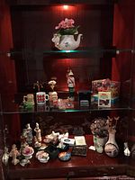 Photo of wooden display cabinet with porcelain/ceramic figurines, teapot planter, mugs, soldier figurine, and small collectibles.