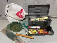 Full fishing tackle box with the lid open, showing various fishing gear, lures, and accessories inside the compartments. A green fishing net with handle, a white plastic bag with mosquito netting, and a wooden handled item beside the box.