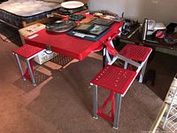 Full view of the folding picnic table set up with 4 attached red perforated seats and red tabletop, shown indoors with various items on tabletop.