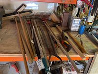 Photo showing multiple wooden-handled garden tools including rakes, axe, pitchfork, and pruning tools on a wooden table in a garage area.