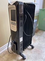 Angled side view of the black oil-filled radiator heater showing its radiating fins, wheels for mobility, and power cord wrapped around the handle.