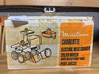 Front side of the original Moulinex Charlotte box showing illustrations of the meat grinder, salad maker, ice crusher cone, and fruit juicer attachments with orange and black text.