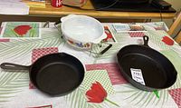 Shows all three items in lot: 8 inch and 10 inch cast iron fry pans and white ceramic casserole dish with stand on table with floral tablecloth.