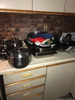 Overview of piles of kitchen cookware items including pots, pans, roasting pans, colanders, and kitchen cloths on countertop.