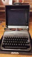 Front view of a gray Remington Rand typewriter in its open hard carrying case showing the keyboard and brand name.