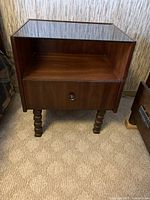 Front view of single wooden side table showing glass top, open shelf, drawer with metal knob, and turned wooden legs.