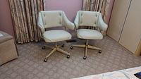Two off-white vinyl office chairs with teal piping and tufted backs on metal four-legged bases with ball casters, side by side on carpet.