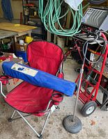 Photo showing one red folding arm chair fully open with a blue carry bag containing the second folding chair placed on top, alongside the work light on its adjustable stand with weighted round base.